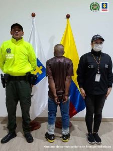 En la foto aparecen de pie, de espaldas, con los brazos atrás y esposado, Carlos Alberto Sinisterra Sinisterra. Viste camisa de manga corta color café oscuro y jean azul. Al lado derecho de él hay un servidor del CTI de la Fiscalía. Al lado izquierdo un uniformado de la Policía Nacional. Detrás de ellos y frente al imputado están las banderas de Colombia y Fiscalía.