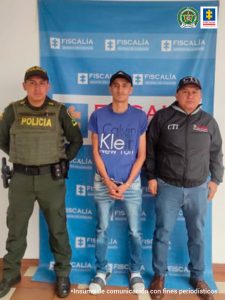 En la foto aparece Carlos Alonso Lemos López, con los brazos adelante y esposado; vestido con camiseta azul de manga corta, jean azul desteñido, gorra, medias blancas y pantuflas. A su lado derecho hay un integrante de la Policía Nacional y a su izquierda un servidor del CTI de la Fiscalía. Detrás de ellos hay un pendón institucional de la Fiscalía.