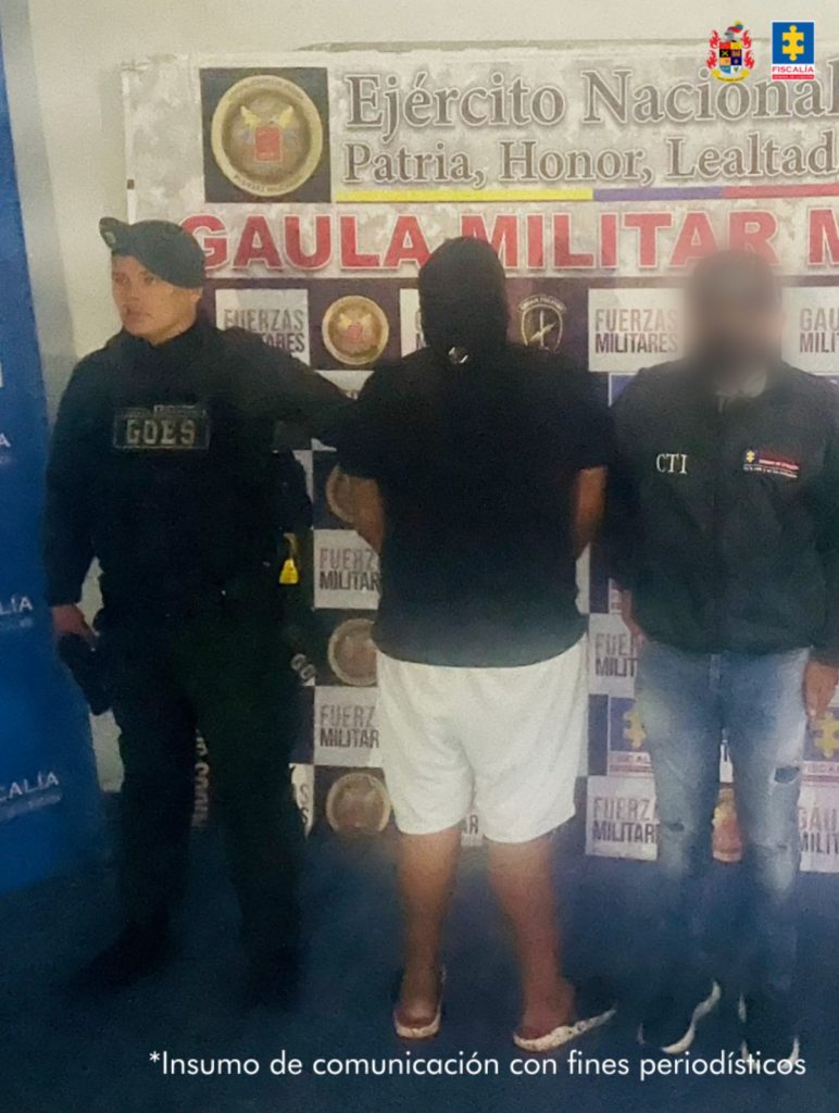 Capturado de frente custodiado por un investigador del CTI de la Fiscalía y un militar. Detrás banner del Grupo Gaula Ariari.