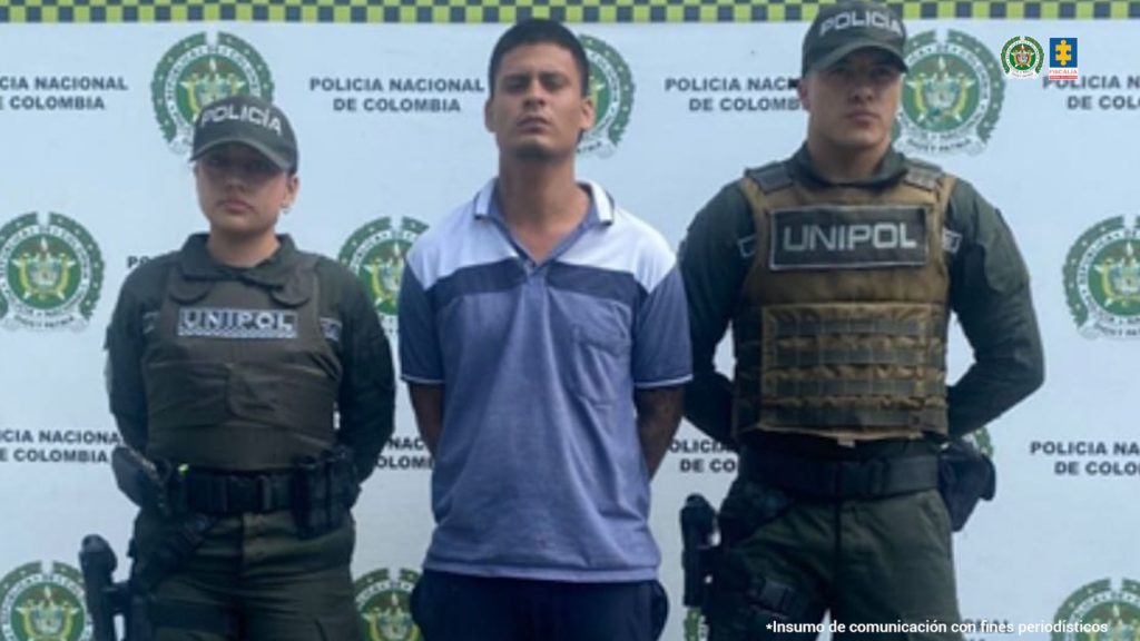 En la fotografía se aprecia de pie y con los brazos atrás a Carlos Andrés Bullas Parra, alias Bullas. Viste camisa de cuello y manga corta, de color azul con franja blanca en la parte del cuello y hombros; asimismo, pantalón azul. A sus extremos hay uniformados de la Policía Nacional. Delante de ellos hay una superficie de color verde y sobre ella hay un letrero que dice: ‘Orden judicial’. Detrás de ellos hay un pendón institucional de la Policía.