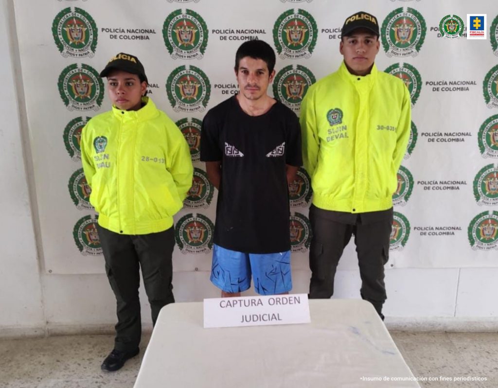 En la foto aparece de pie y con los brazos atrás, Carlos Andrés García Herrera. Esta persona viste camisa negra y pantaloneta azul. A lado y lado de él hay uniformados de la Policía Nacional. Delante de ellos hay una mesa blanca con un letrero sobre ella que dice: captura con orden judicial. Detrás de ellos hay un pendón institucional de la Policía.