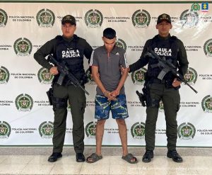 El imputado se encuentra en custodia por dos uniformados del Gaula de la Policía Nacional. El capturado tiene una pantaloneta y un buzo gris. Detrás se observa un backing de la Policía.