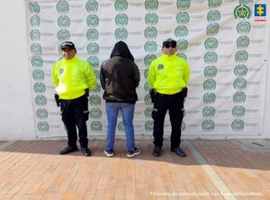 Se visualiza al capturado de espaldas junto a dos uniformados de la Policía Nacional. Detrás de ellos el banner que identifica a la Policía Nacional