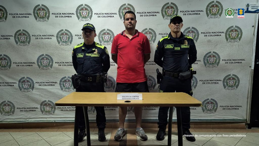 Se observa al capturado de frente y a cada costado un funcionario de policía nacional que lo custodia. Atrás, un pendón de Policía Nacional. Adelante una mesa con el aviso violencia contra servidor público.