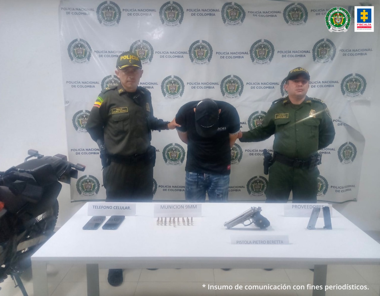 Aparece el hoy imputado, vestido con camisa oscura y jean; con brazos atrás y agachado; acompañado de dos integrantes de la Policía Nacional a lado y lado. Delante de él, hay una mesa con elementos incautados como dos celulares, una pistola, munición y dos proveedores. Asimismo, se ve parte de una motocicleta, igualmente incautada.