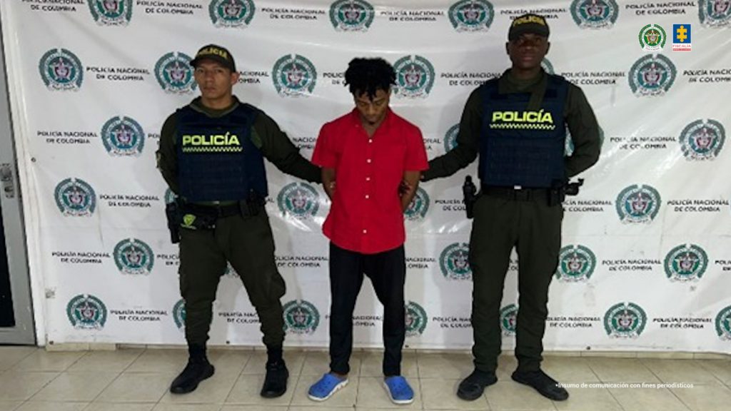 En la fotografía se aprecia de pie y con los brazos atrás a Carlos Manuel Quiñónez. Vista camisa de manga corta de color rojo, pantalón negro y zapatillas azules. A sus extremos hay uniformados de la Policía Nacional. Detrás de ellos hay un pendón institucional de la Policía.