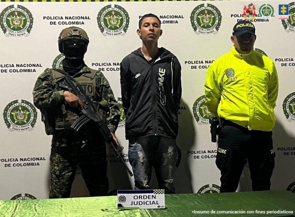 En la fotografía se aprecia de pie y con los brazos atrás a Carlos Manuel Rengifo Gordillo, alias Gato. Viste buso de sudadera negro con estampado vertical al lado izquierdo de él; jean azul degastado. A su extremo izquierdo hay un uniformado de la Policía Nacional. A su derecha un uniformado del Ejército. Delante de ellos hay una superficie de color verde y sobre ella un letrero que dice: ‘Orden judicial’. Detrás de ellos hay un pendón institucional de la Policía.