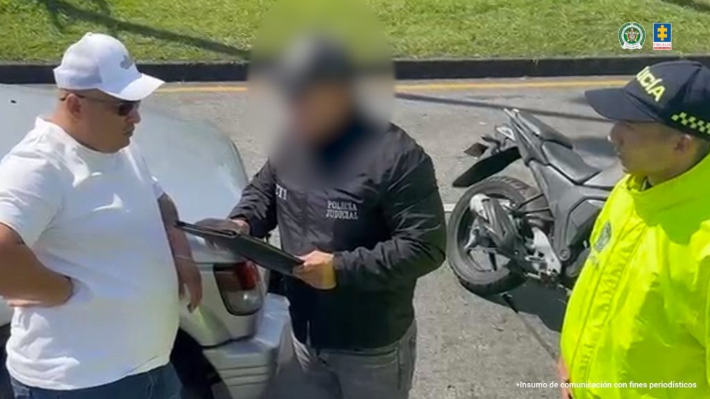 Se observa al capturado en vía pública mientras le son leídos sus derechos por parte de un funcionario del CTI. atrás del capturado un vehículo. De frente al capturado, un funcionario de Policía Nacional