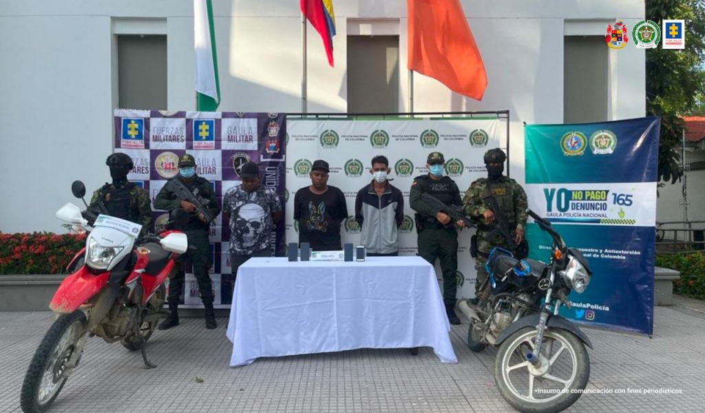 En la imagen están los 3 procesados con un servidor de la Policía y otro del Ejército Nacional a cada lado y con pendones de estas instituciones al fondo.