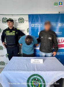  Se visualiza al capturado junto a un uniformado de la Policía Nacional y un investigador del CTI. Detrás de ellos los banners que identifican a la Policía Nacional y a la Fiscalía General de la Nación
