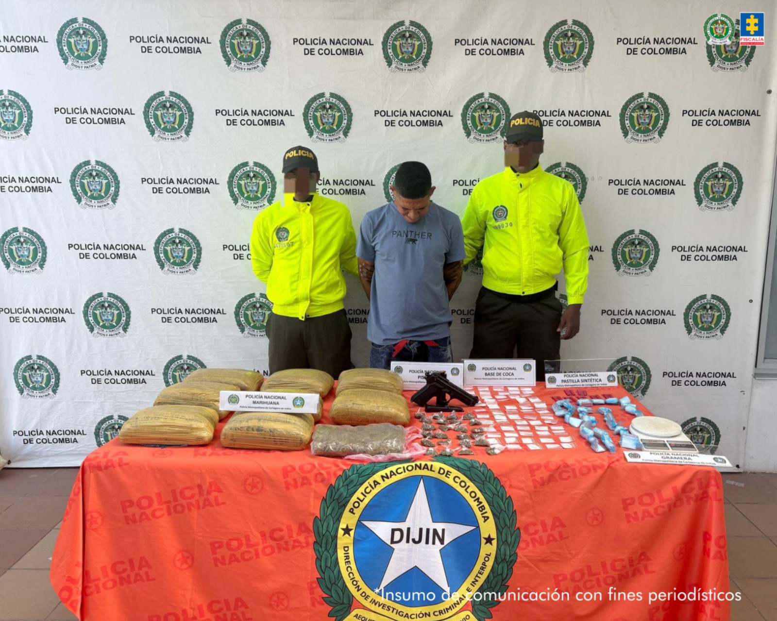 Texto alternativo: En la fotografía aparece un hombre moreno de pie, con la cabeza agachada, brazos atrás esposado, delgado, vestido con una camiseta azul, custodiado por dos uniformados de la Policía Nacional. Delante de ellos hay una mesa con la droga y un arma de fuego.