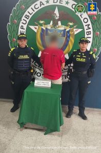 Se observa al capturado de espaldas y a cada costado un funcionario de Policía Nacional.  Atrás un Escudo de Colombia y Policía Nacional.  Al frente una mesa con el cartel CAPTURA POR ORDEN JUDICIAL.