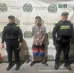 En la foto aparece el capturado custodiado por dos uniformados de la Policía Nacional. En la foto aparece detrás un backing de la Policía Nacional. El capturado tiene unas pantalonetas con bordes de colores y un buso con muñecos animados.