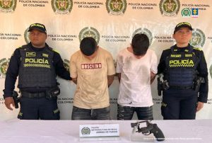 En la foto aparecen los dos capturados custodiado por dos uniformados de la Policía Nacional. En la foto aparece detrás un backing de la Policía Nacional y una mesa con un arma de fuego.