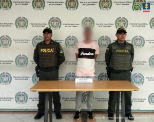 Se observa al capturado de frente y a cada costado un funcionario de Policía Nacional.  Atrás un pendón de Policía Nacional.  Al frente una mesa con el cartel VIOLENCIA INTRAFAMILIAR.