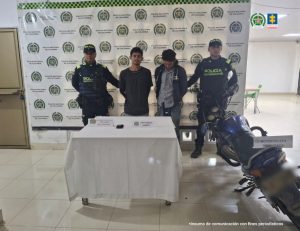 En la foto aparecen los dos capturados custodiado por dos uniformados de la Policía Nacional. En la foto aparece detrás un backing de la Policía Nacional y una mesa con un celular y también aparece una motocicleta.