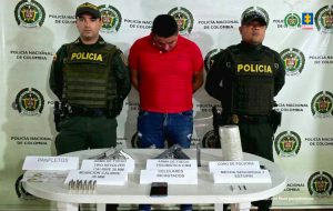 En la foto aparece el capturado de frente y custodiado por dos uniformados de la Policía Nacional. En la foto también aparece un backing de la Policía Nacional y el armamento, munición, panfleto y pólvora incautada.
