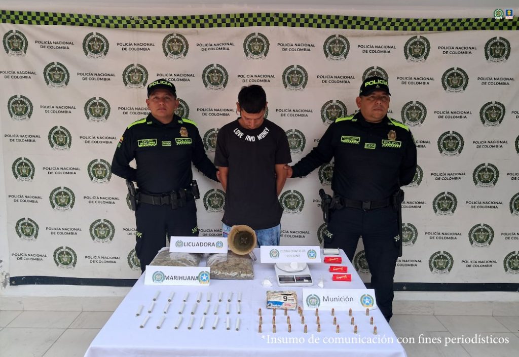 En la foto aparece el capturado de frente y custodiado por dos uniformados de la Policía Nacional.  En la foto también aparece de fondo un backing de la Policía Nacional y el material incautado durante la orden de registro y allanamiento.