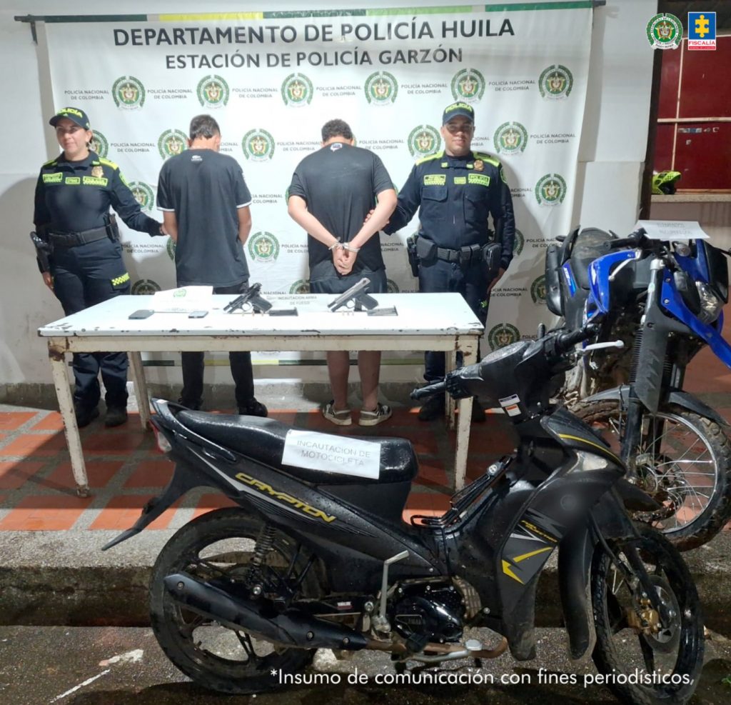 En la foto se observa a los dos capturados de espalda custodiados por dos uniformados de la Policía Nacional. Detrás hay un backing de la Policía Nacional. En la foto se observa la moto recuperada y las armas incautadas durante el operativo.