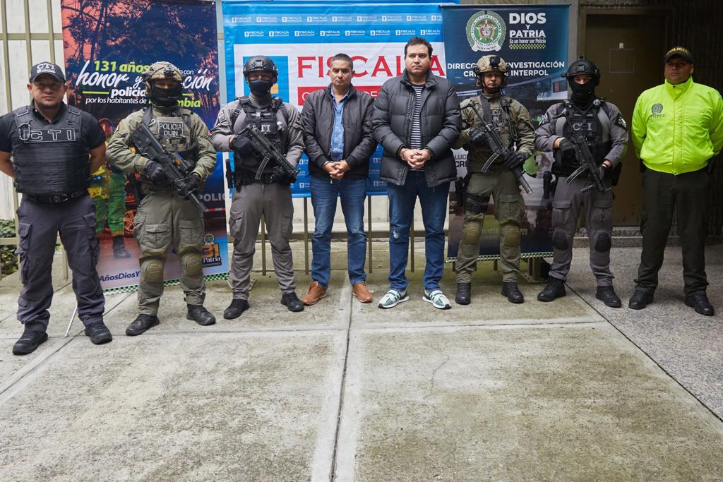 En la imagen aparecen nueve personas. Las dos personas del centro, vestidas de civil son los hermanos Andrés Felipe y Ramón Emilio Pérez Hoyos, por su posible participación en el homicidio del fiscal de Paraguay Marcelo Daniel Pecci Albertini. A los costados están uniformados de la policía quienes vigilan a los procesados. Al fondo se aprecia una pancarta donde aparecen los logos de policía y Fiscalía. Estas personas están en las instalaciones de la Fiscalía General de la Nación.