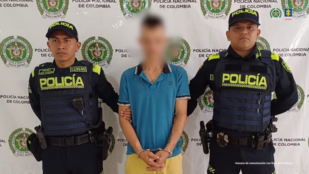 En la foto se observa al capturado custodiado por dos uniformados de la Policía Nacional. El capturado tiene una camiseta azul y una pantaloneta de color amarilla.