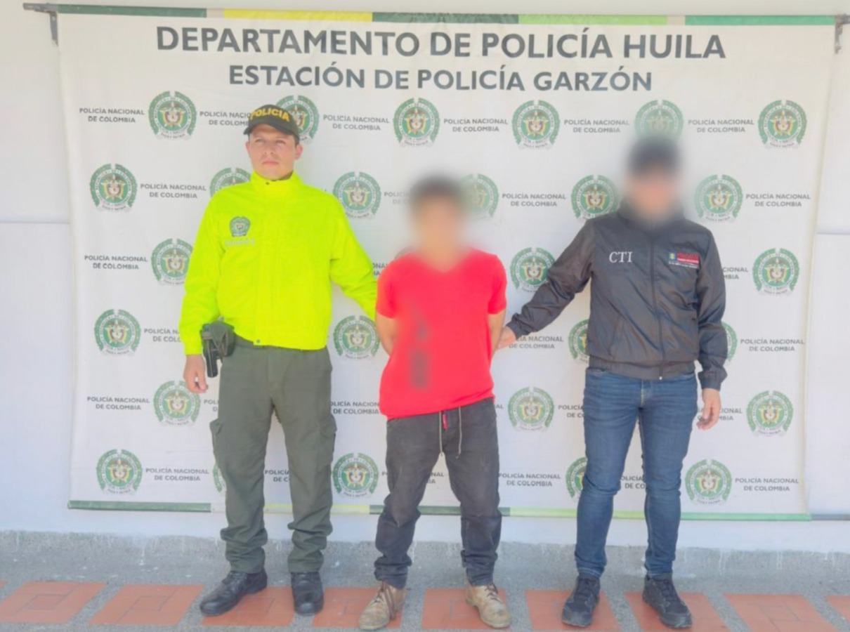 El capturado se encuentra de frente y tiene una camiseta color roja. Se encuentra custodiado por dos uniformados de la Policía Nacional.