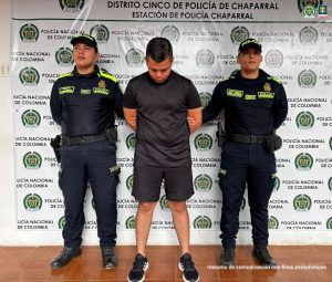 En la imagen se ve una persona detenida bajo custodia de dos integrantes de la Policía Nacional. Detrás suyo un backing institucional.