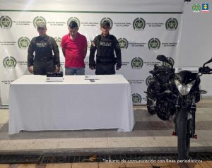  En la foto aparecen el capturado de frente y custodiados por dos uniformados del Gaula de la Policía Nacional. Detrás aparece un backing de Gaula y también aparece una mesa con el material incautado. 