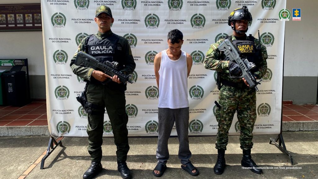 En la foto se observa a uno de los capturados, custodiado por dos uniformados del Gaula de la Policía Nacional. También se observa un backing de la Policía Nacional.