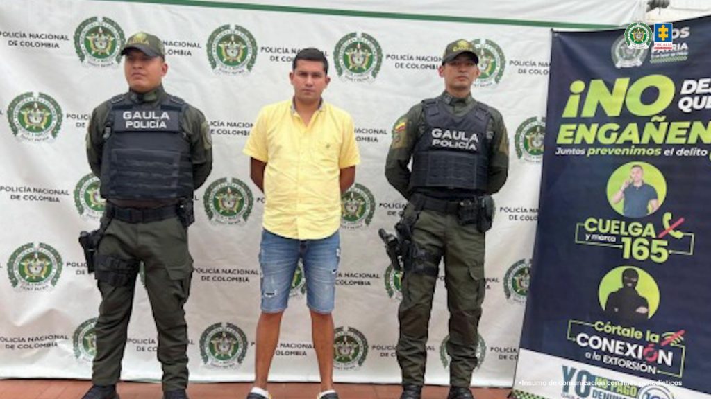 En la foto se observa al capturado custodiado por dos uniformados del Gaula de la Policía Nacional. Detrás hay un backing de la Policía Nacional.
