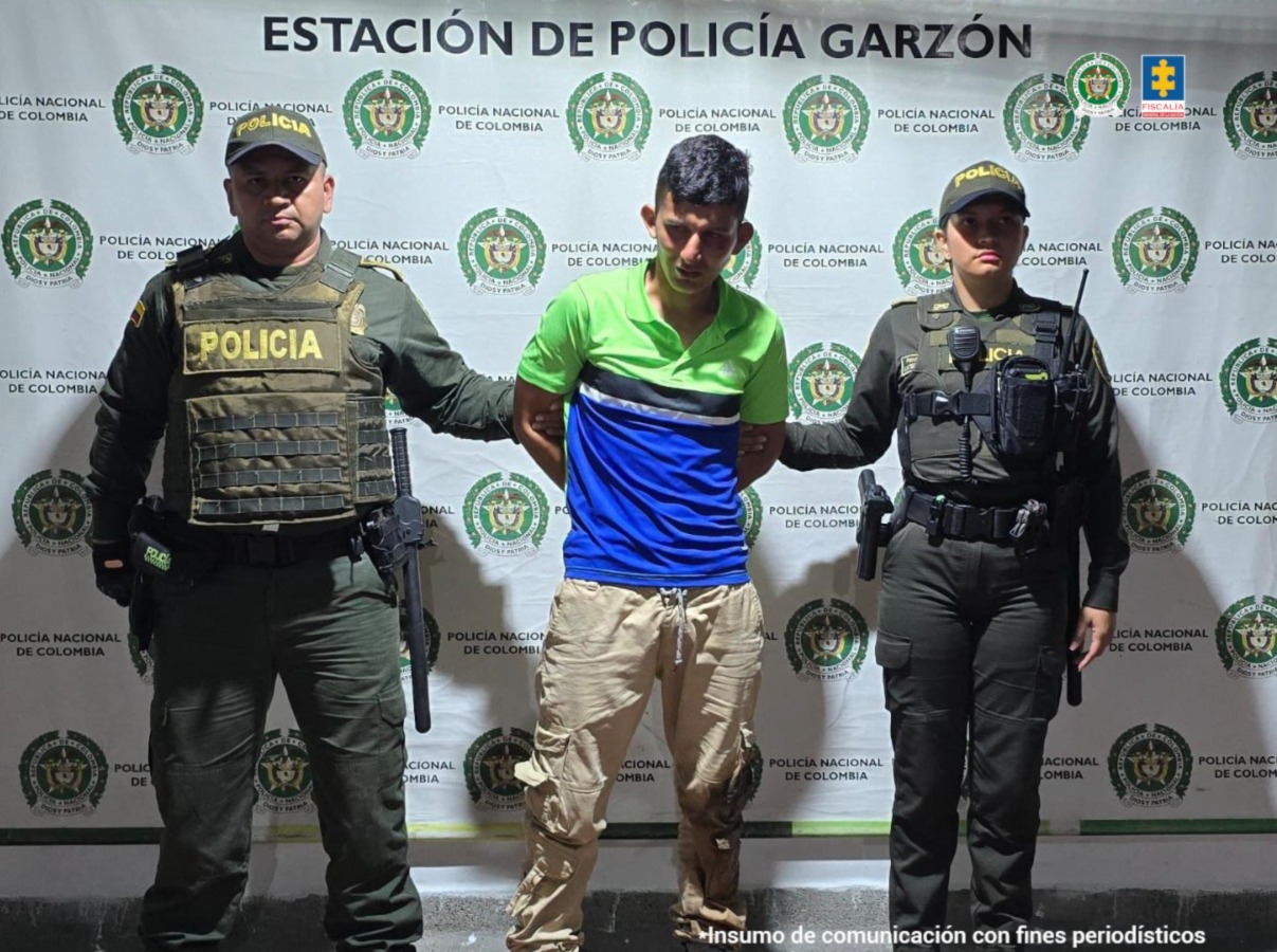 El capturado se encuentra de frente y custodiado por dos uniformados de la Policía Nacional. En la foto se ve de fondo un backing de la Policía Nacional.