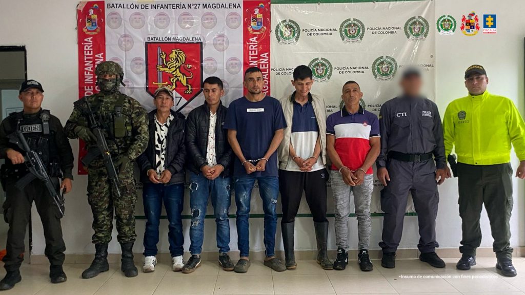 Los capturados se encuentran de frente y custodiados por un servidor del Cuerpo Técnico de Investigación CTI, un uniformado del Ejército y GOES de la Policía Nacional. Detrás se observa un backing de la Policía y Ejército Nacional. 