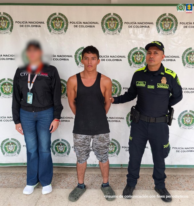 En la foto aparece el capturado de frente y custodiado por un servidor del Cuerpo Técnico de Investigación CTI y un uniformado de la Policía Nacional. En la foto aparece de fondo un backing de la Policía Nacional.