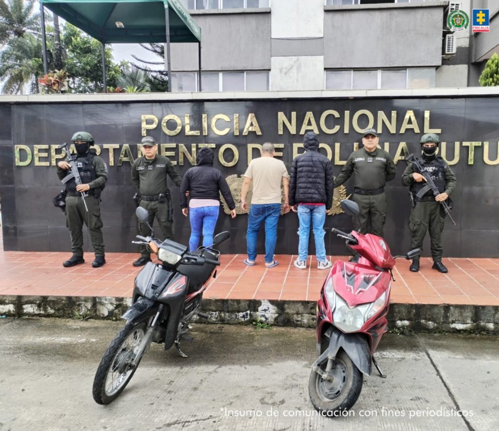 En la foto aparece los capturados de espalda y custodiados por dos uniformados de la Policía Nacional.  En la foto también aparece de fondo un backing de la Policía Nacional y las dos motos incautadas