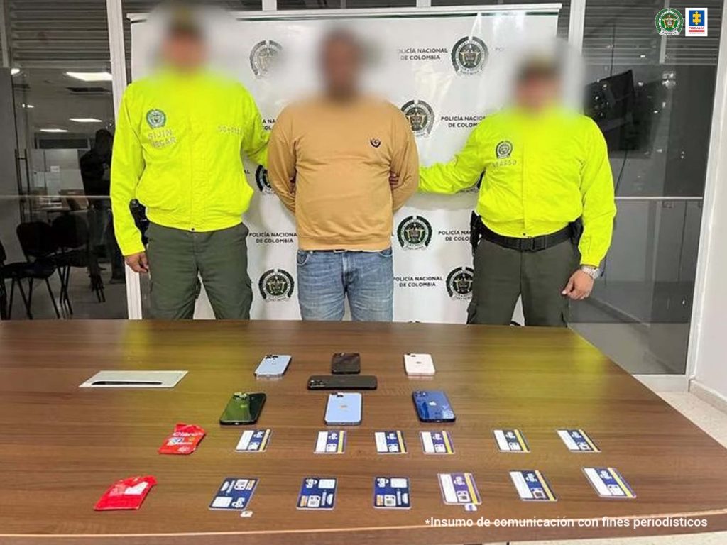 En la imagen se observa a un hombre de buso amarillo y jean azul claro custodiado por dos agentes de la Policía Nacional. Delante de él se identifica una mesa con celulares y tarjetas de celulares.