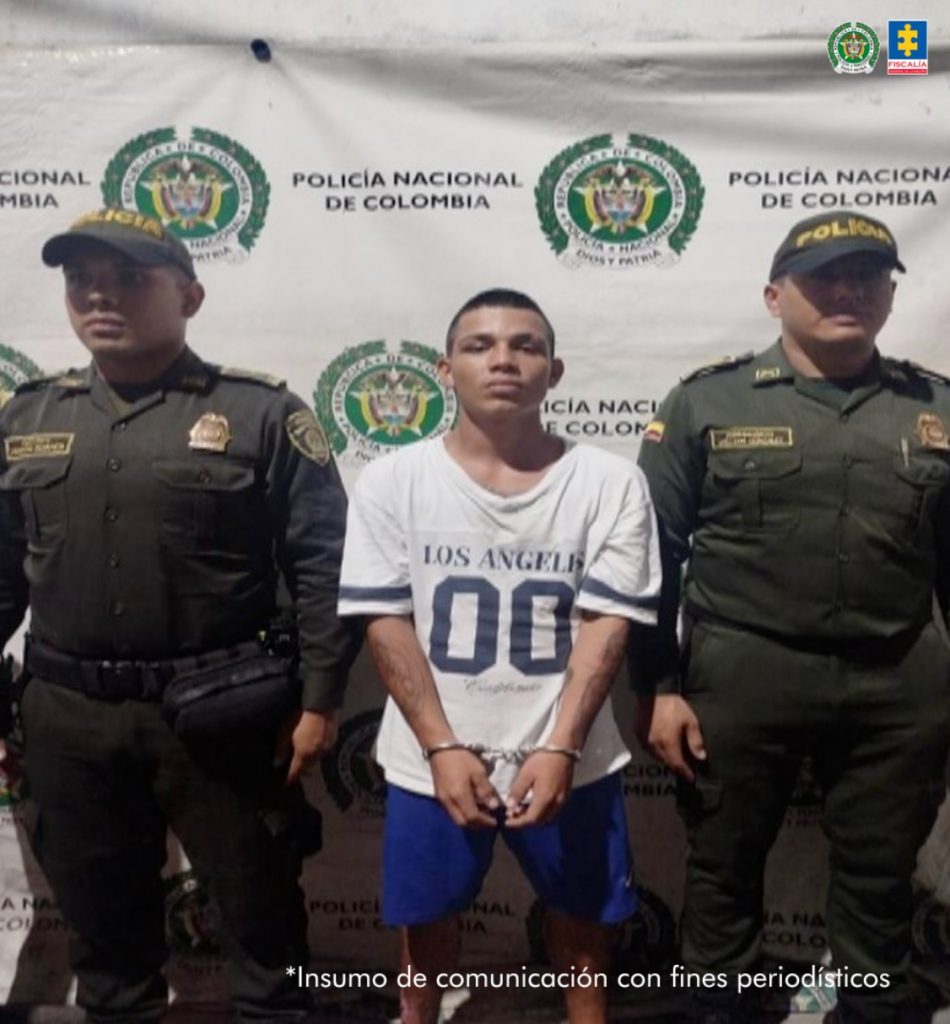 El capturado se encuentra de frente y custodiado por dos uniformados de la Policía Nacional. Detrás se observa un backing de la Policía Nacional.