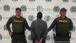 En la foto aparece el capturado de espalda y custodiado por dos uniformados de la Policía Nacional. En la foto aparece de fondo un backing de la Policía Nacional.