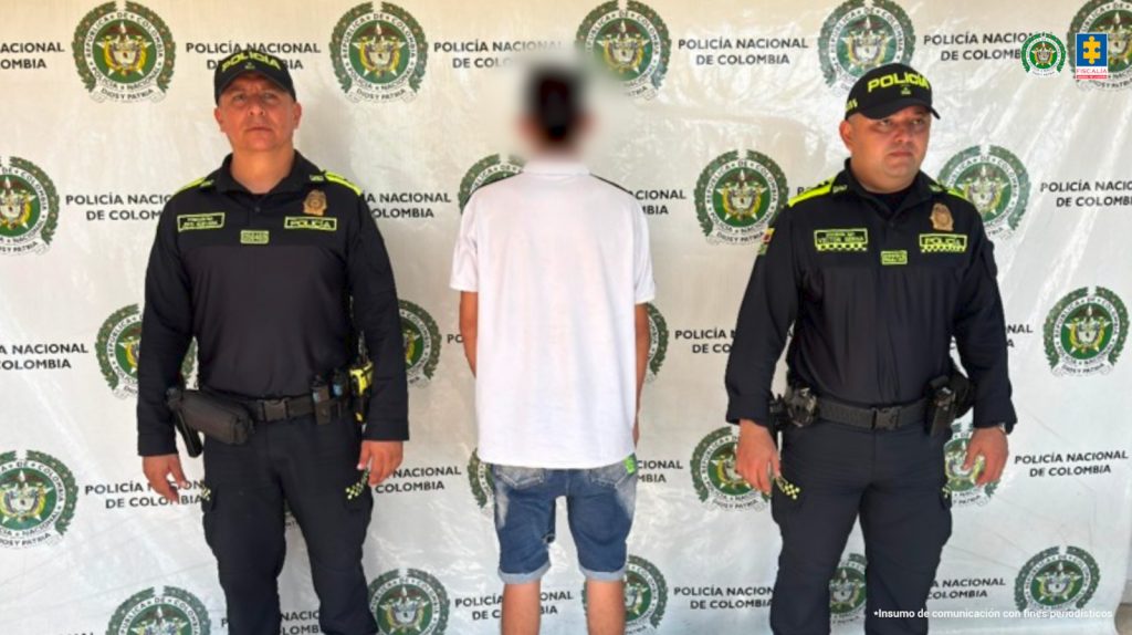 En la foto aparece el menor aprehendido de espalda y custodiado por dos uniformados de la Policía Nacional. El menor tiene puesta una bermuda de jean y un buso blanco. De fondo aparece un backing de la Policía Nacional.