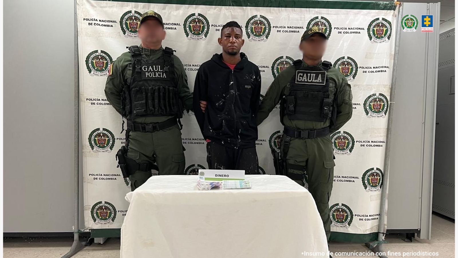 En la imagen está un hombre moreno, alto de pie, vestido con buzo y pantalón negro, tiene los brazos atrás esposado y es de cabellos crespos cortos negros. Se encuentra custodiado por dos uniformados del Gaula de la Policía Nacional. 