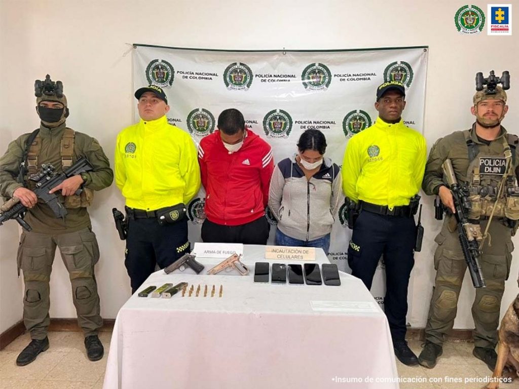 En la foto aparecen de pie, los brazos atrás y la cabeza agachada César Eduardo Montoya, alias ‘Negro’ o ‘César’, y Katerine Cárdenas Ruiz. El hombre usa tapabocas y viste camisa roja de manga corta con rayas blancas diagonales en las mangas y pantalón negro. La mujer usa tapabocas y viste buzo color beage de manga larga y jean azul claro. A lado y lado de ellos hay uniformados de la Policía Nacional. Delante de ellos hay una mesa con mantel blanco y sobre ella hay dos armas de fuego, tres proveedores, munición y cuatro celulares. Asimismo, hay dos letreros que dicen: ‘Armas de fuego’ e ‘Incautación de celulares’. Detrás de ellos hay un pendón institucional de la Policía.