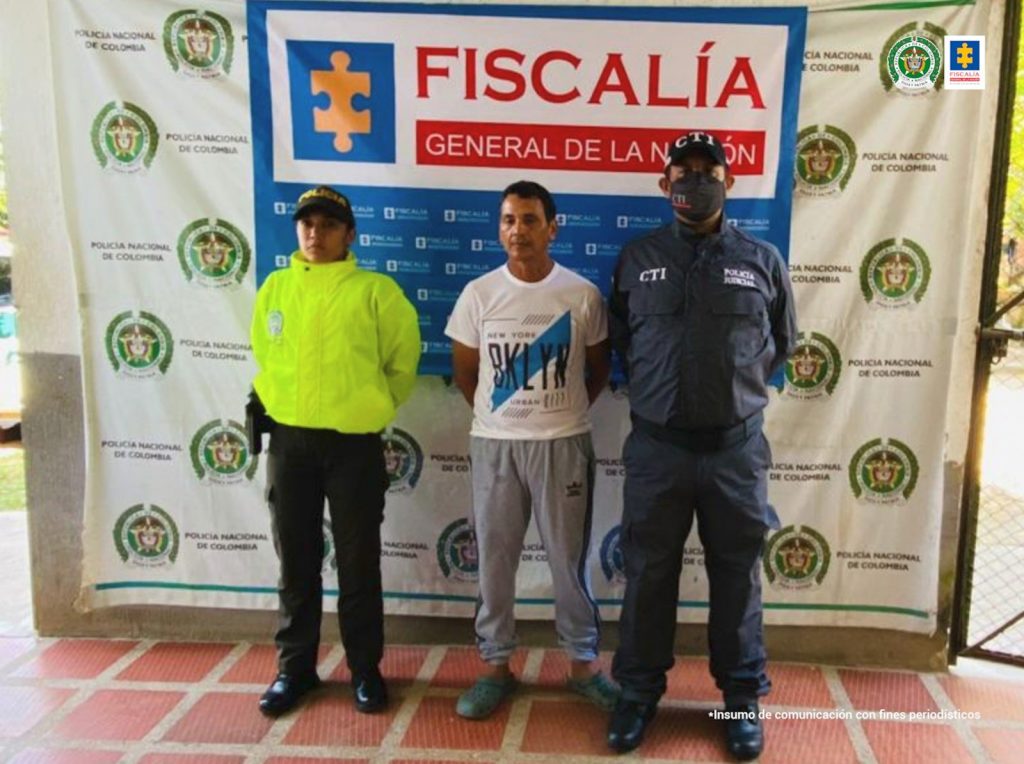 está el capturado de frente esposado manos atrás, custodiado por un investigador del CTI de la Fiscalía y por un Policía. Detrás banner de la Fiscalía y de la Policía Nacional.
