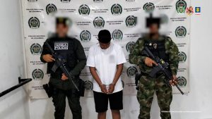 El imputado se encuentra en custodia por un uniformado del Gaula de la Policía y el Ejército Nacional. El capturado tiene un buzo blanco. Detrás se observa un backing de la Policía.