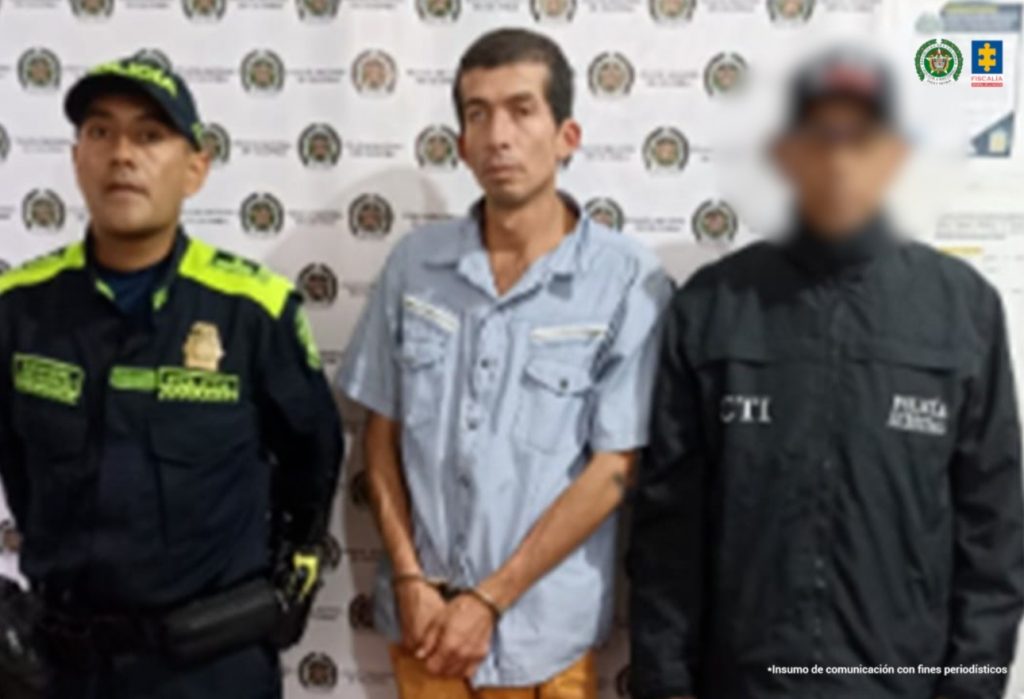 En la fotografía se aprecia de pie, con los brazos adelante y esposado a César Piter Vásquez Erazo. Viste camisa de manga corta, de color azul claro; pantalón de color mostaza, zapatillas negras con zuela blanca. Al lado izquierdo hay un servidor del CTI de la Fiscalía y al lado derecho hay un uniformado de la Policía Nacional. Detrás de ellos hay un pendón institucional de la Policía.