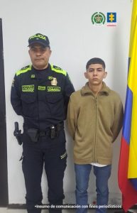 En esta foto se observa un procedimiento de captura oficial en Colombia:A la izquierda aparece un policía uniformado, con gorra que dice “POLICÍA” y con insignias visibles en su uniforme.  A la derecha está un joven con chaqueta beige y pantalón claro, posando junto al uniformado con las manos hacia atrás.  En el fondo, sobre la pared blanca, se distinguen los logos de la Policía Nacional de Colombia y la Fiscalía General de la Nación.  A la derecha de la imagen también se ve una parte de la bandera de Colombia.  En la parte inferior aparece la leyenda: “Insumo de comunicación con fines periodísticos”.