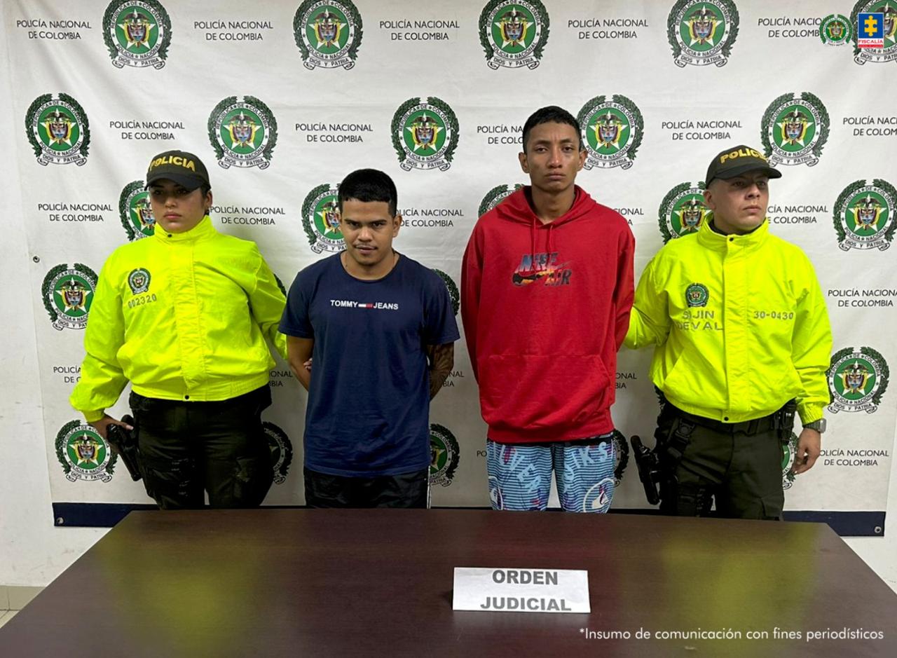 En la imagen aparecen dos hombres capturados, rodeados a cada lado por miembros de la Policía Nacional. Al fondo puede verse un banner de la Policía. Frente a ellos hay una mesa con un letrero que dice "orden judicial".