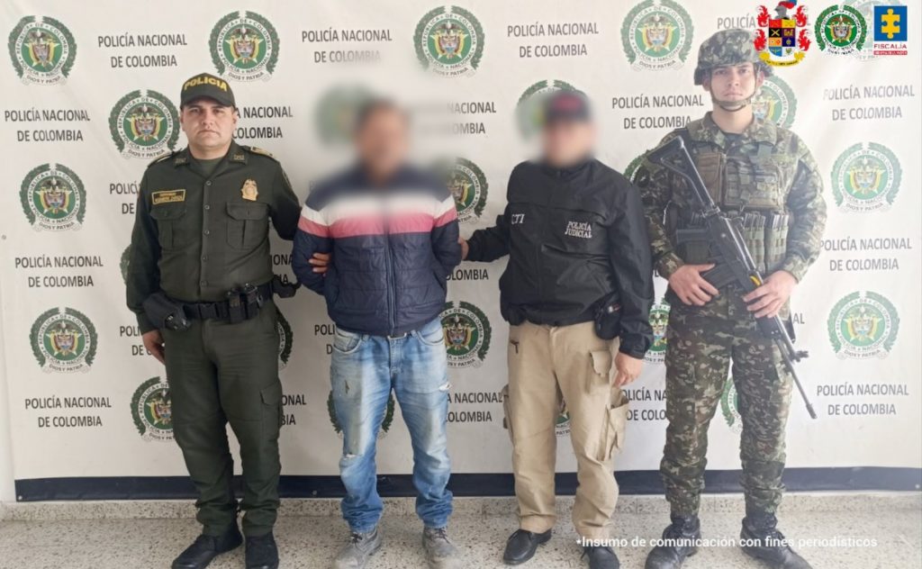 fotografia en donde se ve al capturado en medios de un agente de policia, un funcionario CTI y un soldado del ejercito, atras de ellos baking de la policia