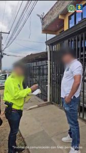 En la foto se observa al uniformado de la Policía Nacional cuando le está leyendo la orden de captura al ciudadano Daniel Andrés Urbano Meneses . El procesado tiene una camiseta de color blanco y un jean. En la foto se observa unas casas de rejas.