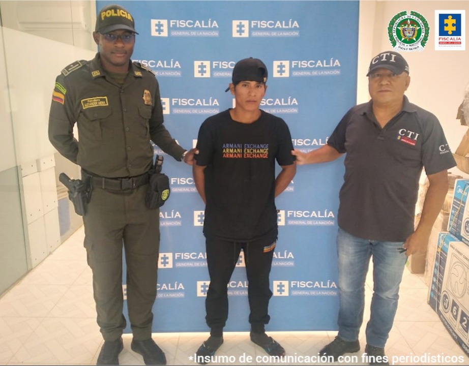 El capturado aparece frente a un banner de la Fiscalía. A cada lado tiene personal del CTI de Fiscalía y de la Policía.