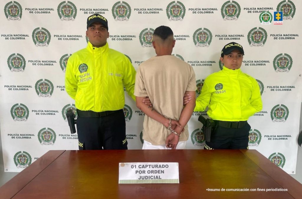 Fotografia en donde se ve al capturado de espaldas custodiado por dos policias, detras de ellos hay un pedon de la policia y delante de ellos uhay una mesa con un letrero