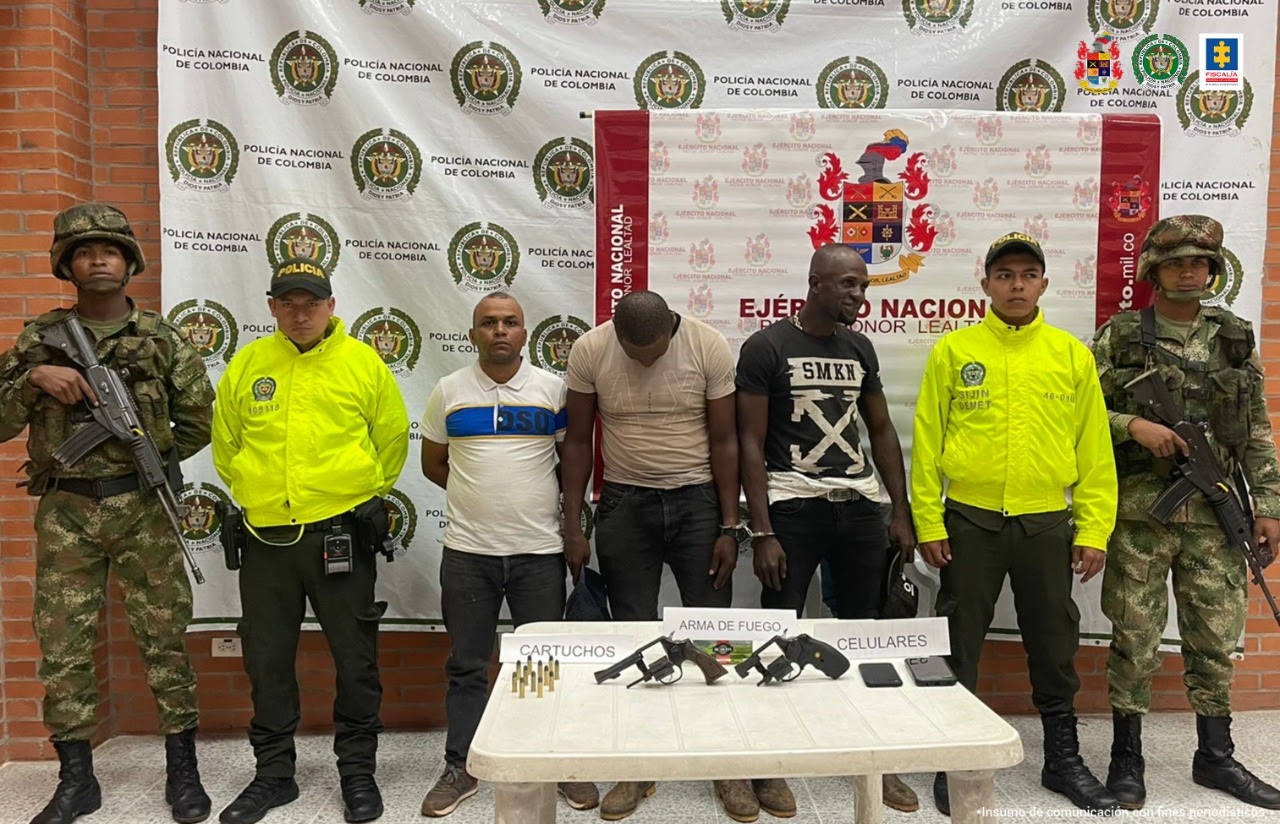En la foto están los tres capturados de frente custodiados por dos uniformados de la Policía y dos militares del Ejército Nacional, detrás de ellos los banner de las dos instituciones y delante de ellos una mesa con el material incautado en las diligencias